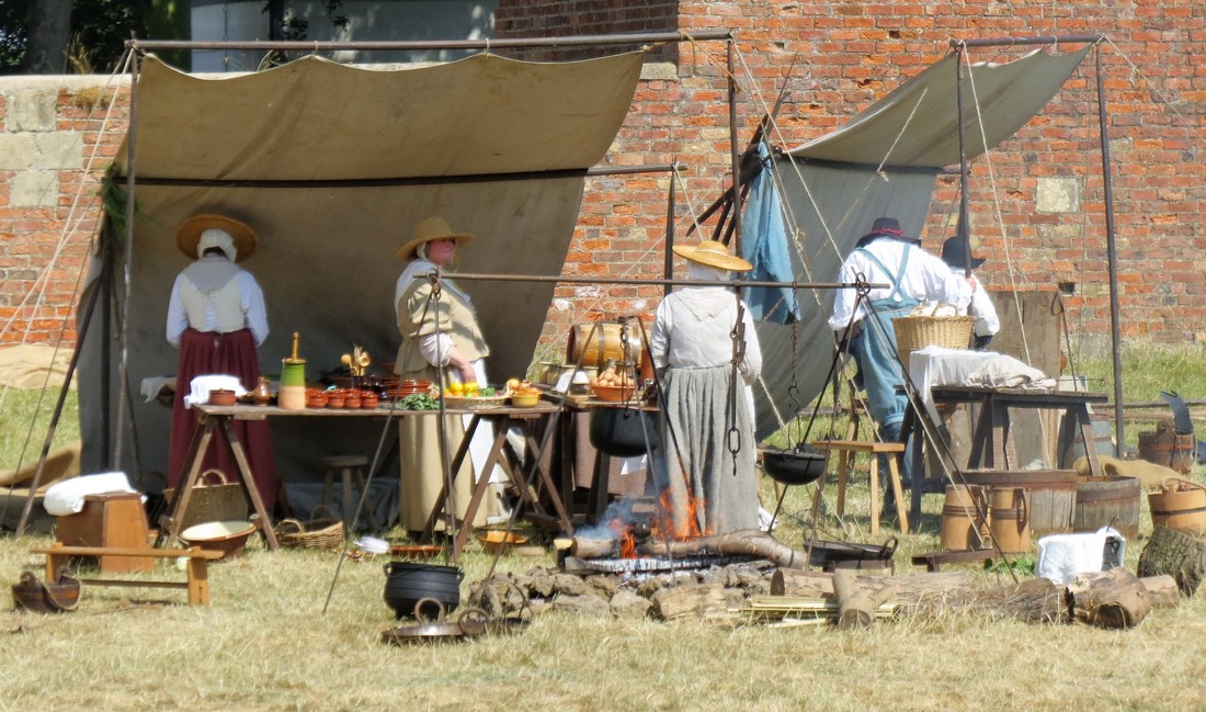 Civilians of Lunsford's stand around the kitchen tent and the metalworking tent. A cauldron hangs over a fire in the foreground.